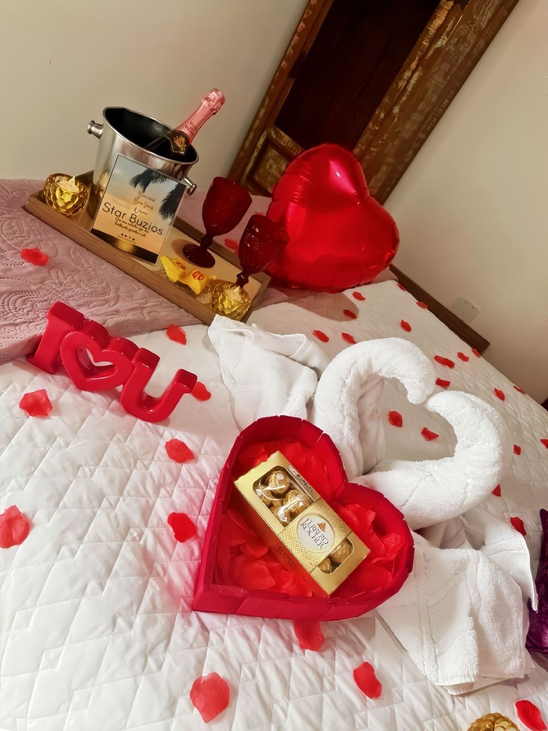 Cama decorada em estilo romântico com corações vermelhos e brancos, rosas e caixas de presente sobre uma colcha acolchoada.
