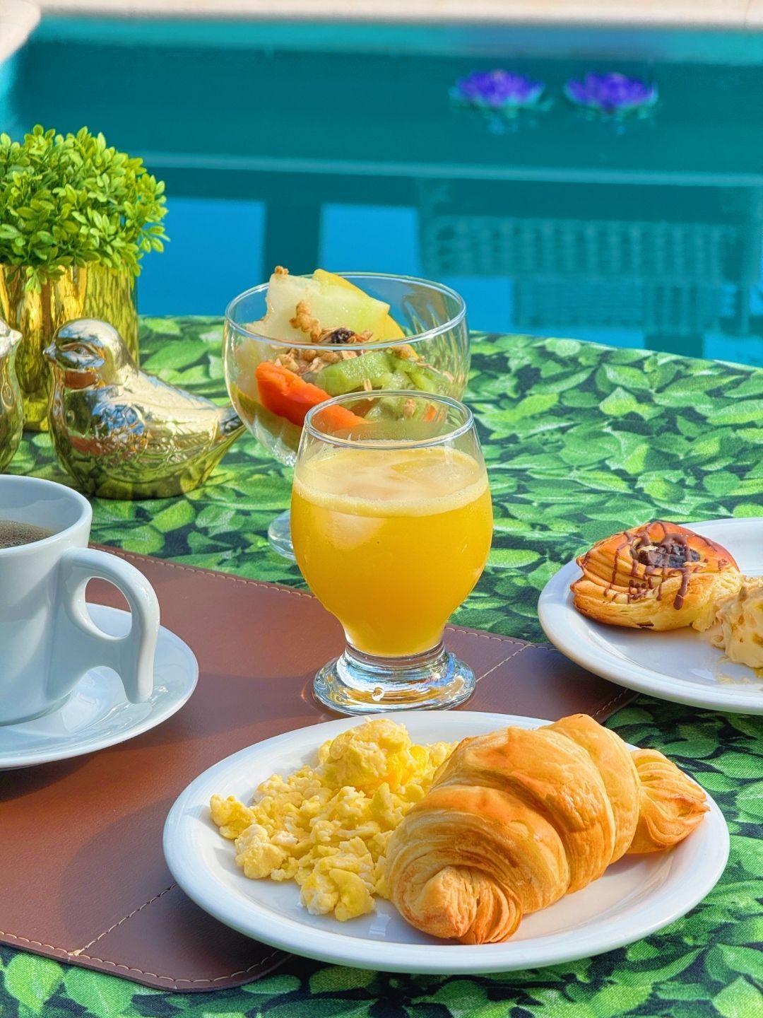 Café da manhã servido em uma mesa à beira da piscina, com café, suco de laranja, croissant, ovos e salada de frutas.