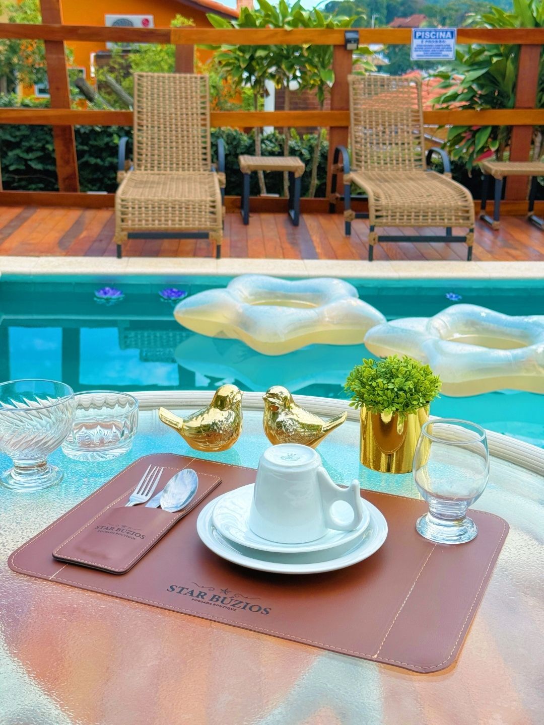 Bandeja de café da manhã à beira da piscina com café, doces e flores, ao lado de uma piscina azul e cadeiras de vime.