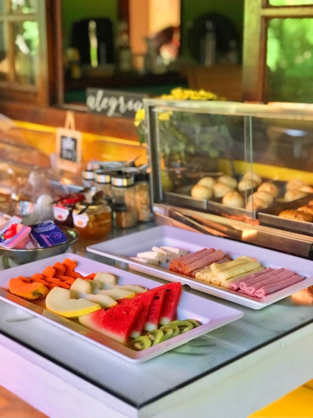 Bandejas de sushi e aperitivos expostas em um balcão de buffet branco, com pinças e uma vitrine refrigerada de vidro atrás.