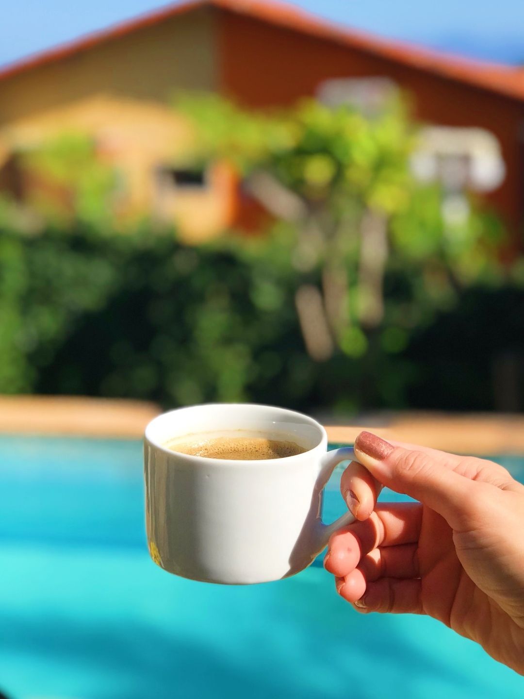 Mão segurando uma xícara de café branca à beira de uma piscina azul brilhante com uma casa desfocada ao fundo.