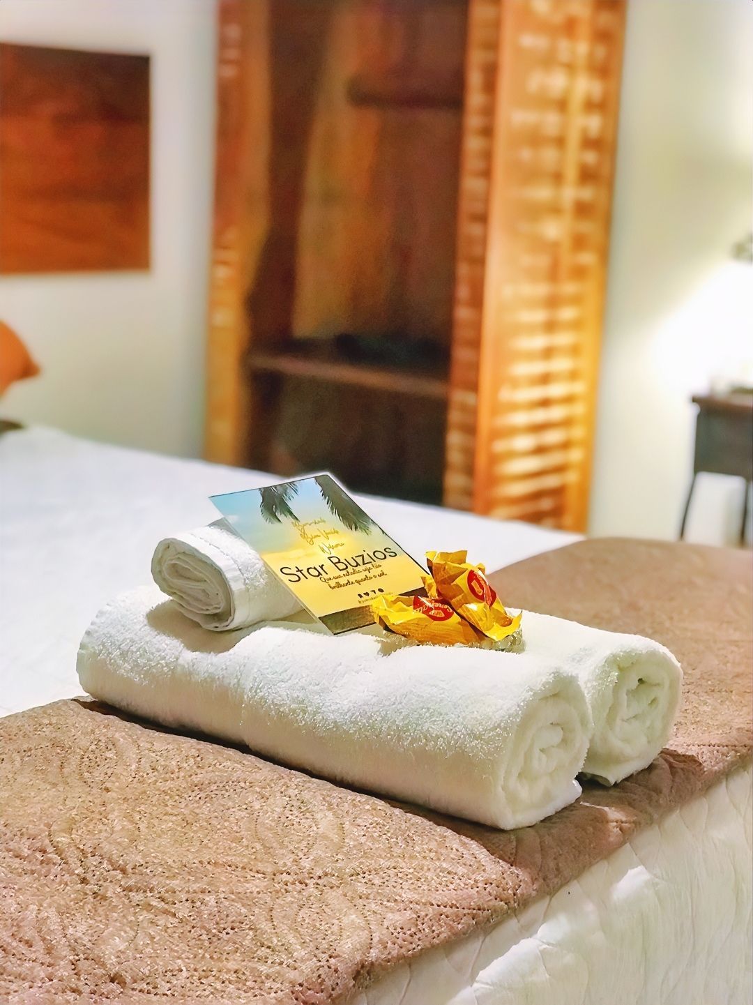 Toalhas brancas enroladas sobre uma cama com uma pequena flor e um folheto em um quarto de hotel.