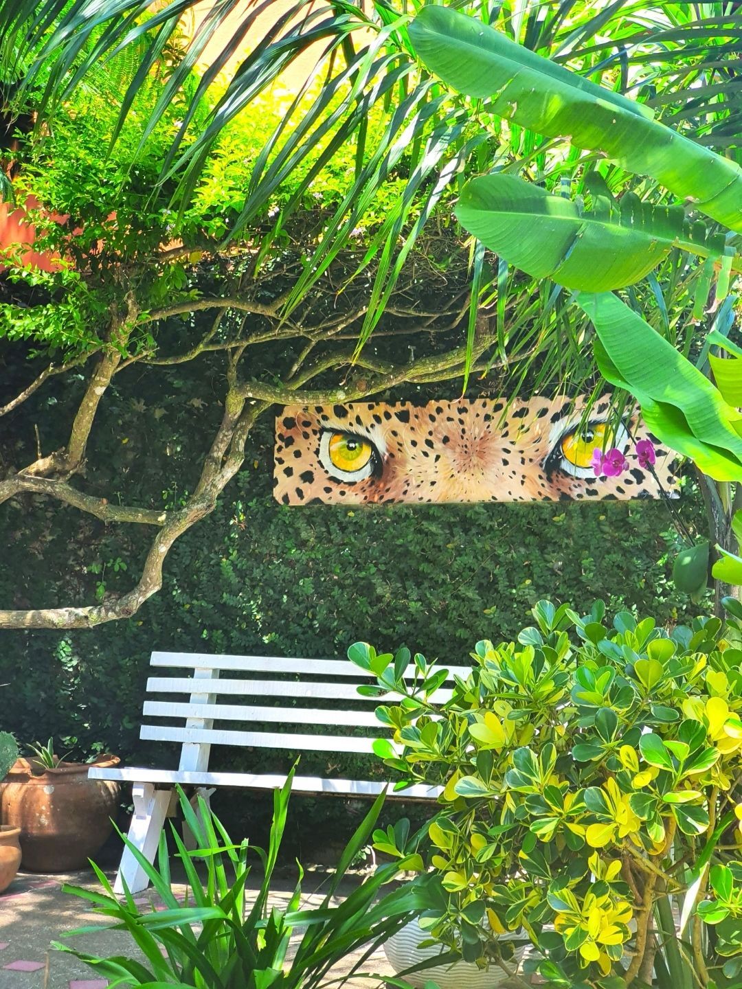 Banco branco em um exuberante jardim tropical com um mural de um leopardo espreitando por entre a vegetação.