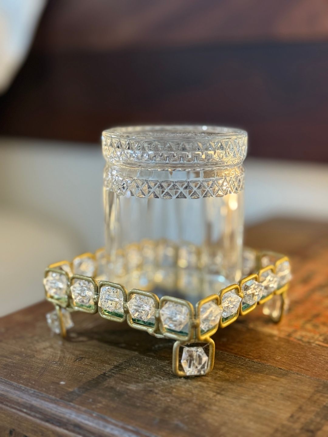 Castiçal de vidro sobre um suporte dourado com detalhes em pedraria, apoiado em uma superfície de madeira.