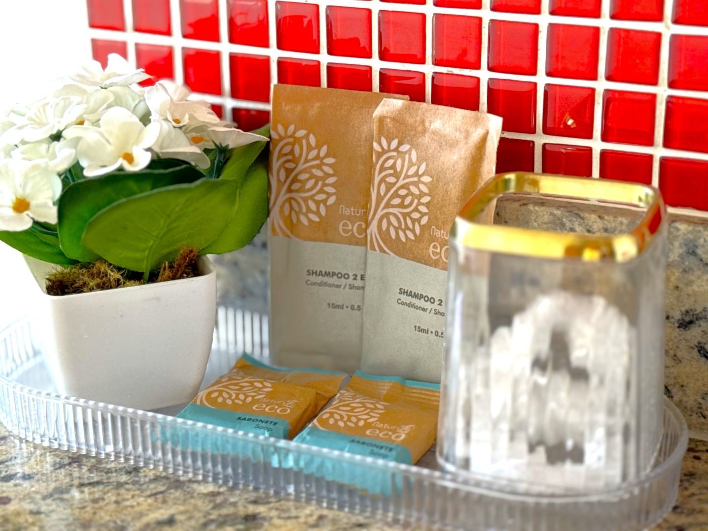 Orquídea branca com itens embalados em dourado e um recipiente de vidro em uma bandeja contra uma parede de azulejos vermelhos.