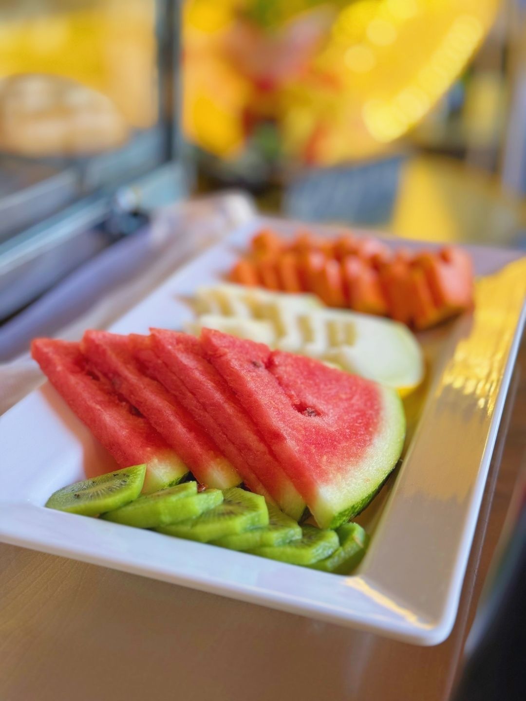 Bandeja com frutas variadas fatiadas, incluindo melancia, kiwi e melão, dispostas em um bufê.