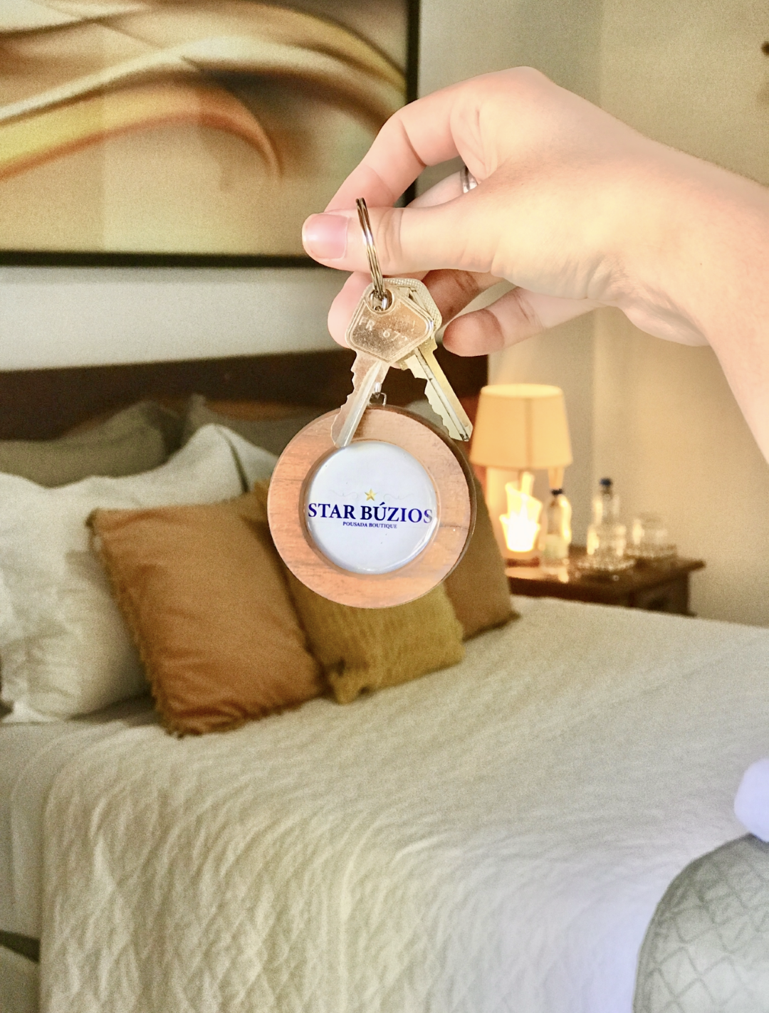 Mão segurando um chaveiro redondo bege sobre uma cama arrumada com travesseiros brancos e bege em um quarto aconchegante.