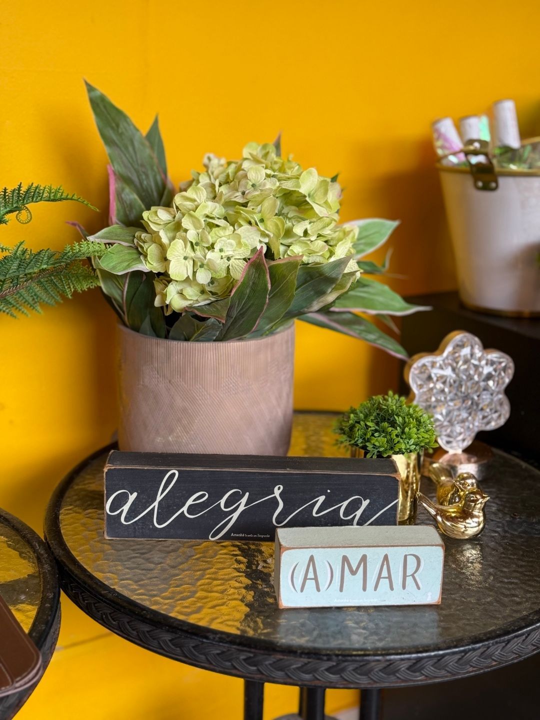 Flores decorativas e placas de lousa sobre uma bandeja redonda preta contra uma parede amarela.