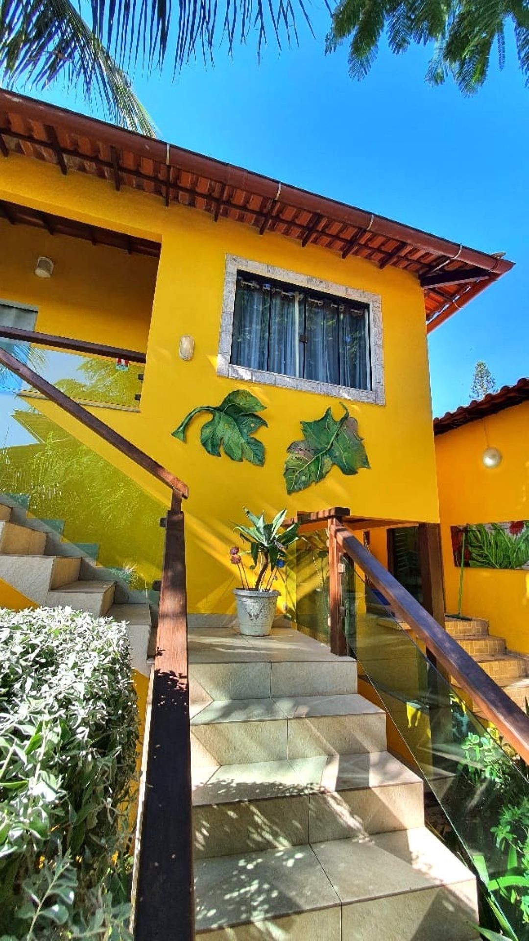 Casa amarela brilhante com degraus de pedra, corrimãos de madeira e murais de palmeiras verdes sob um céu azul.