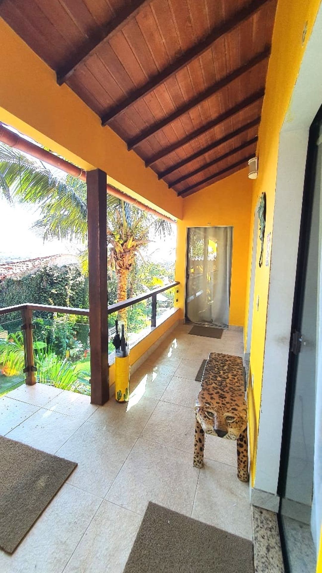 Varanda externa coberta com paredes amarelas, teto de madeira, corrimão e um banco ao lado da porta.