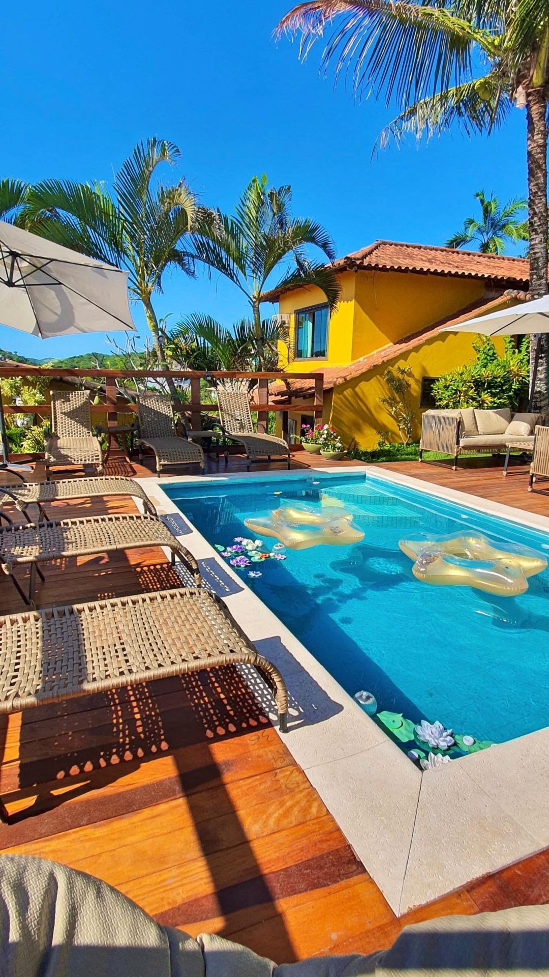Piscina tropical no quintal com espreguiçadeiras, guarda-sóis, palmeiras e uma casa amarela sob um céu azul.