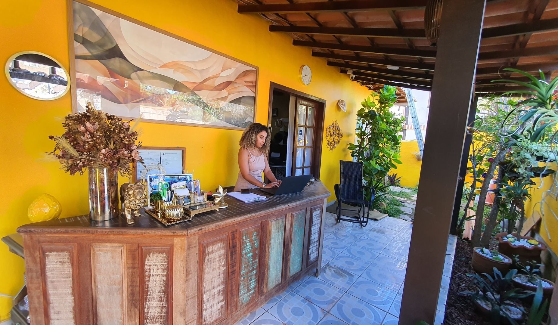 Balcão de recepção estilo resort com balcão de madeira entalhada, paredes amarelas e plantas tropicais sob uma varanda coberta.