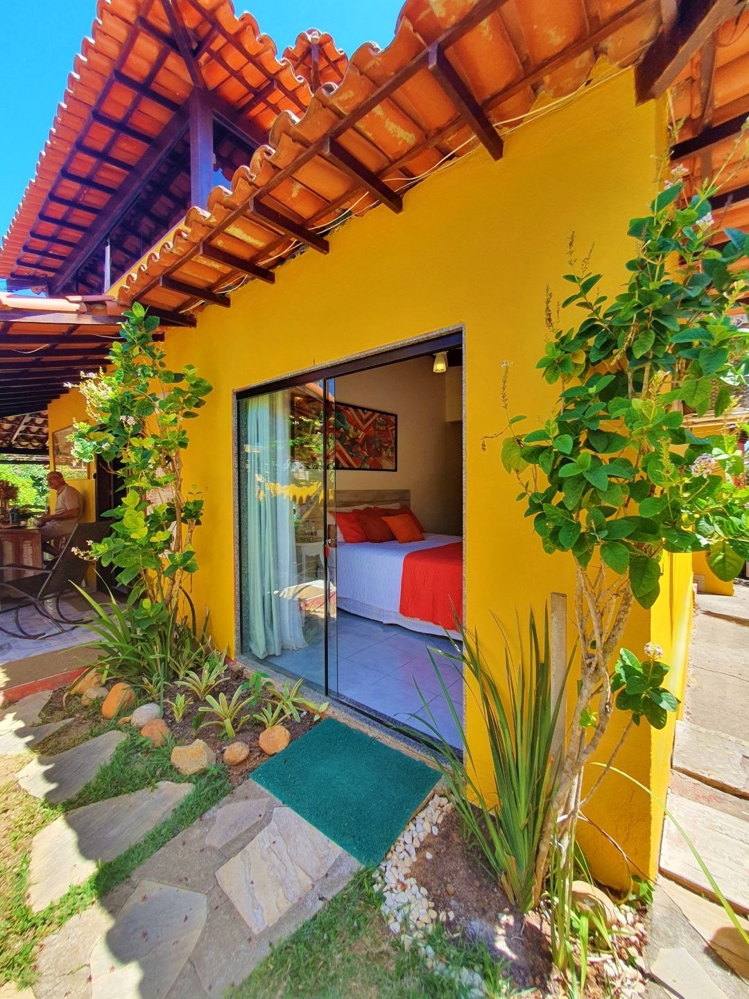 Exterior colorido de chalé amarelo com porta de correr aberta, cama vermelha no interior e exuberantes plantas em vasos junto ao caminho.