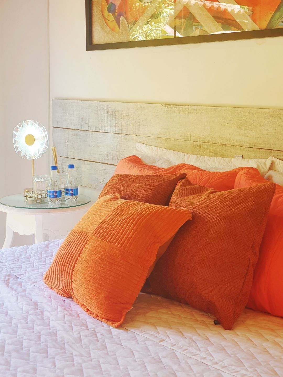 Quarto aconchegante com colcha branca, almofadas laranja, mesa de cabeceira e arte floral na parede.