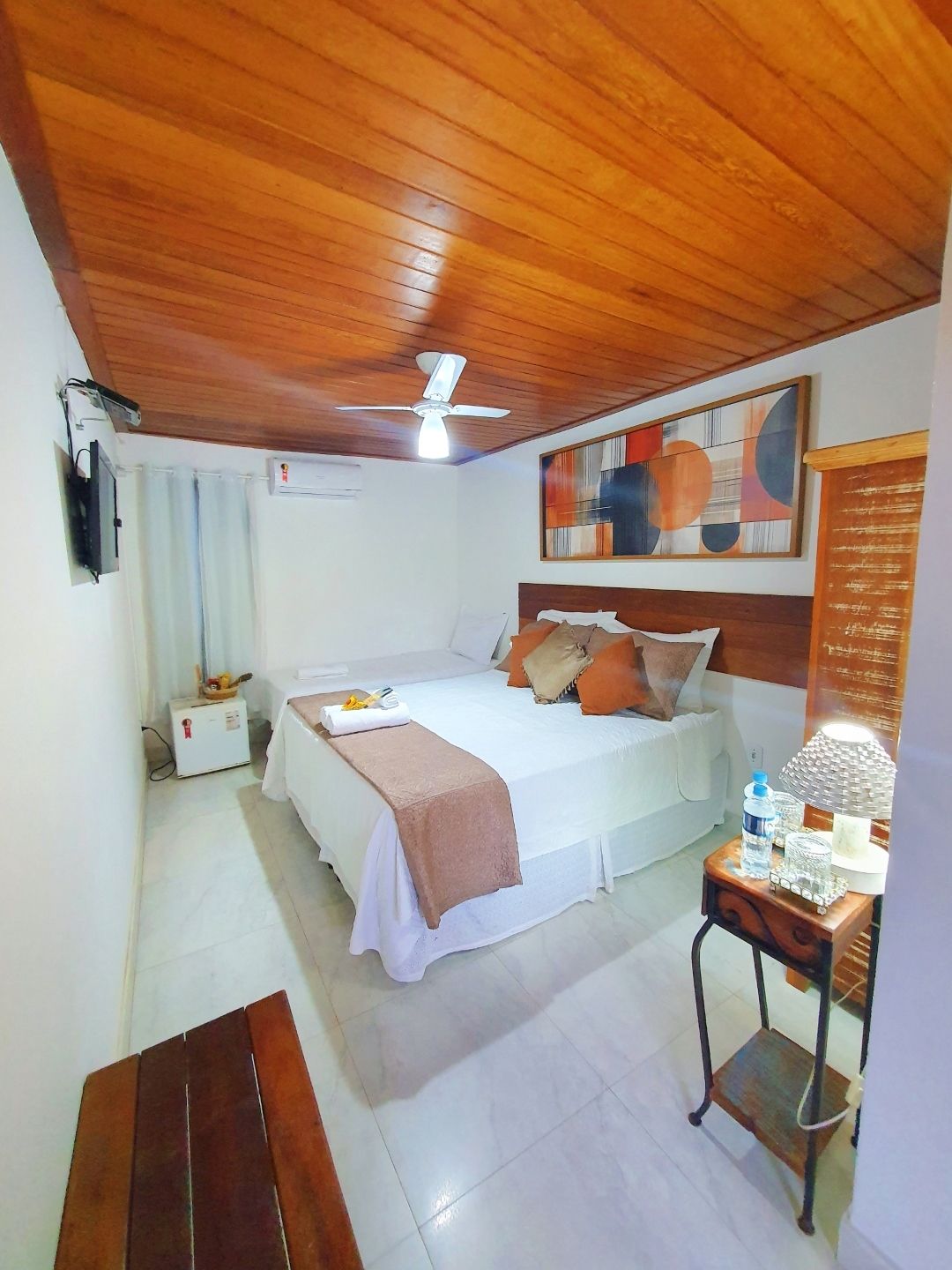 Quarto aconchegante com teto de madeira, roupa de cama branca, detalhes em laranja e arte na parede.