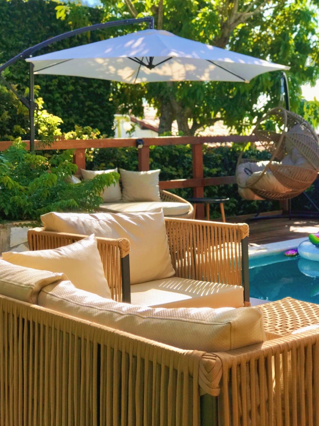 Pátio ensolarado com espreguiçadeiras de vime, guarda-sol branco e vegetação à beira da piscina.