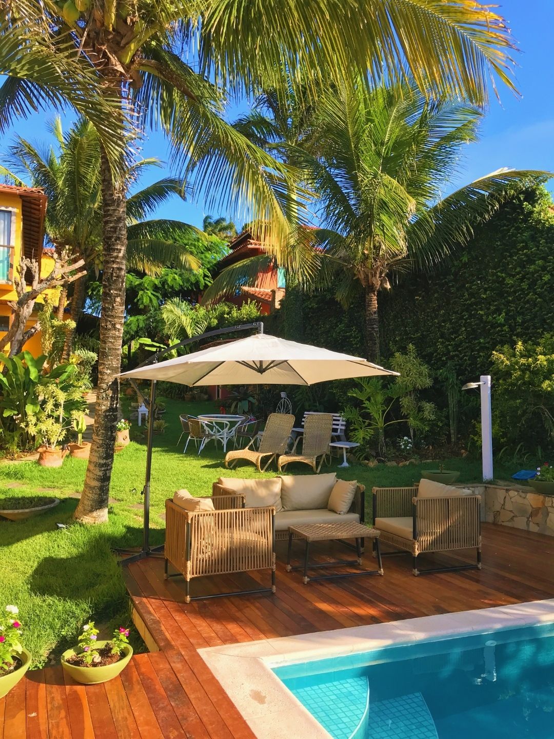 Pátio tropical à beira da piscina com guarda-sol branco, espreguiçadeiras, palmeiras e água turquesa sob um sol radiante.