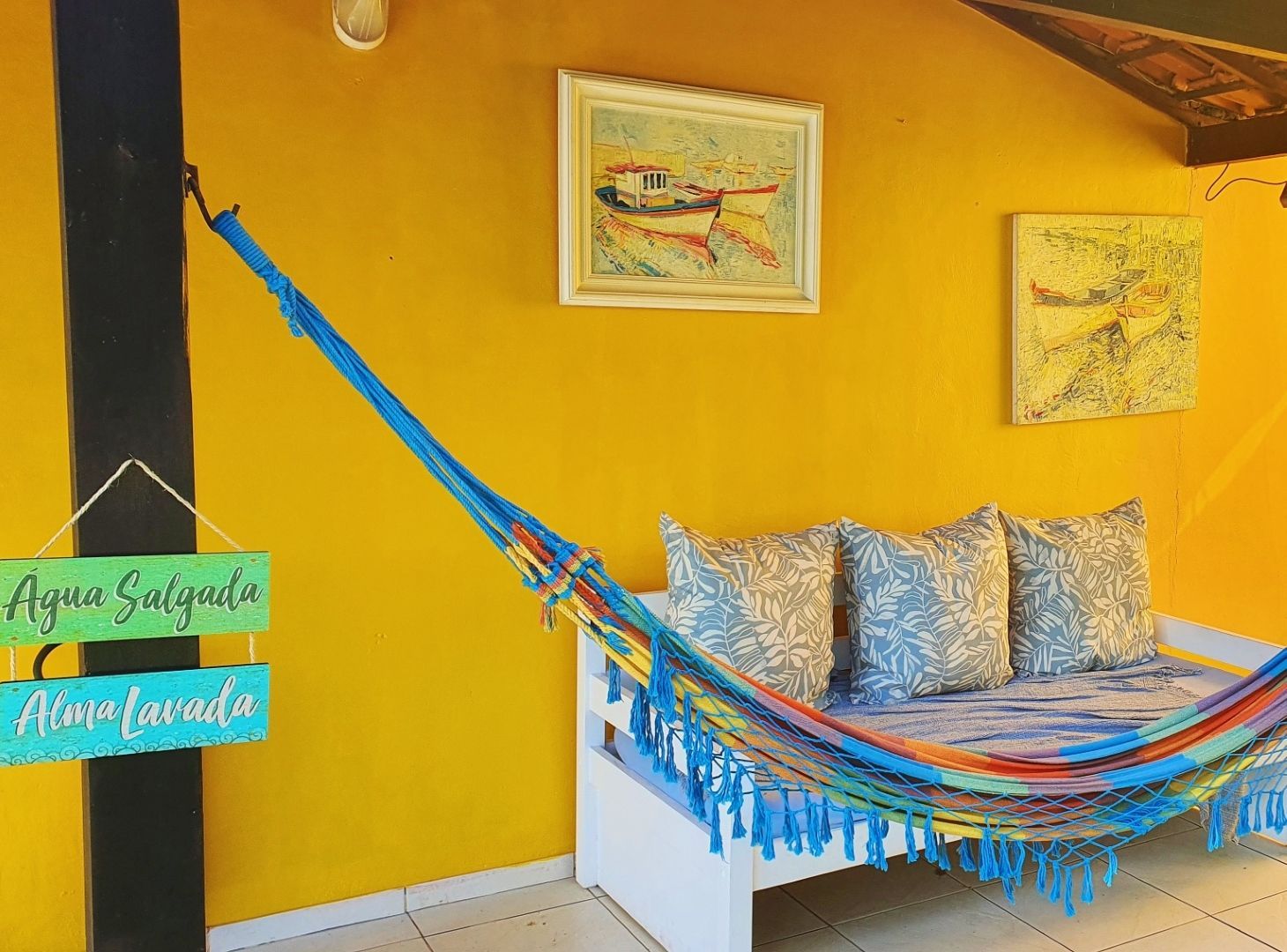 Rede colorida ao lado de uma parede amarela com obras de arte emolduradas e uma pequena placa, em um quarto aconchegante.