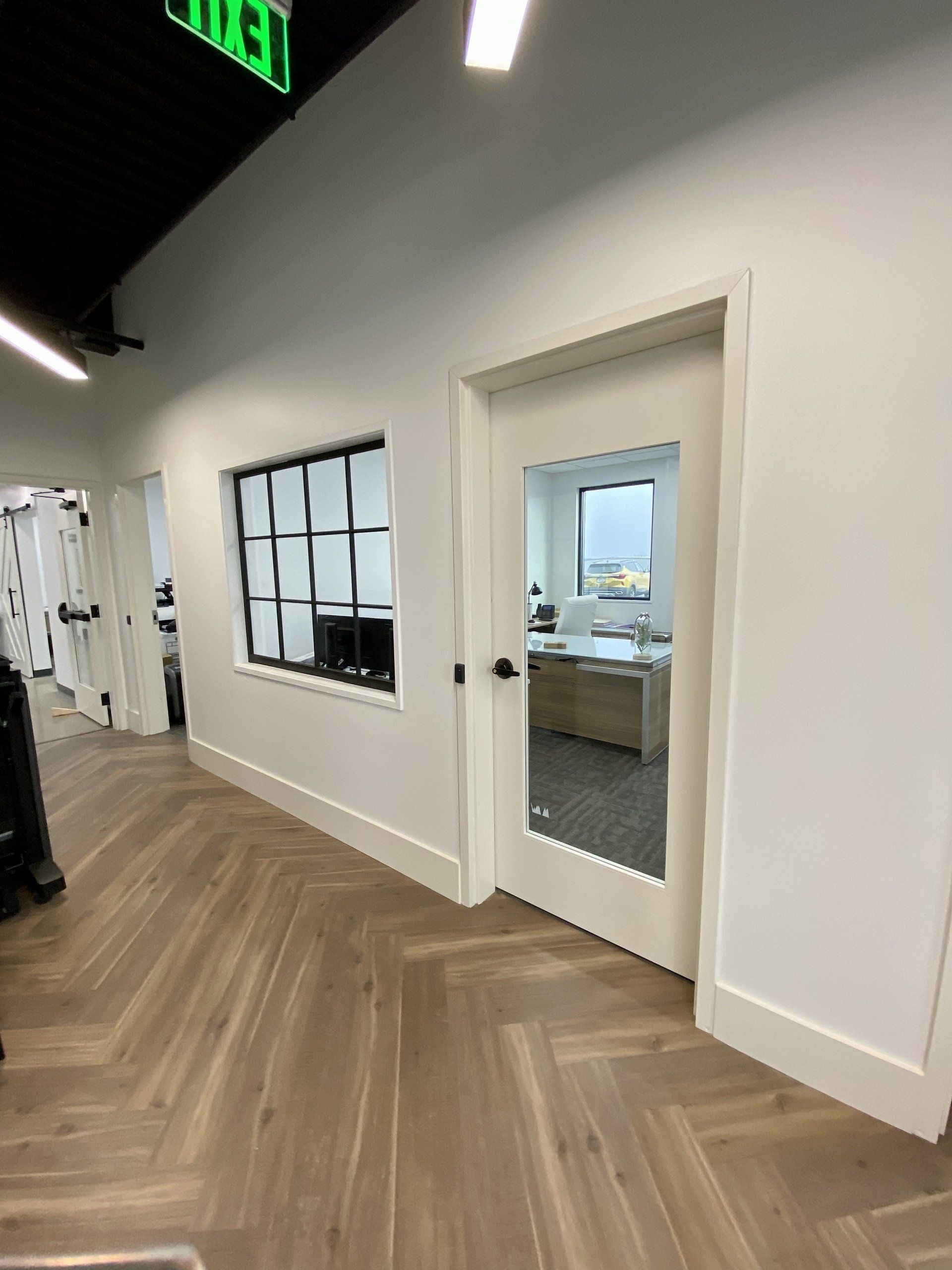 Bright office hallway with white walls, wood floors, and a glass door into a meeting room.