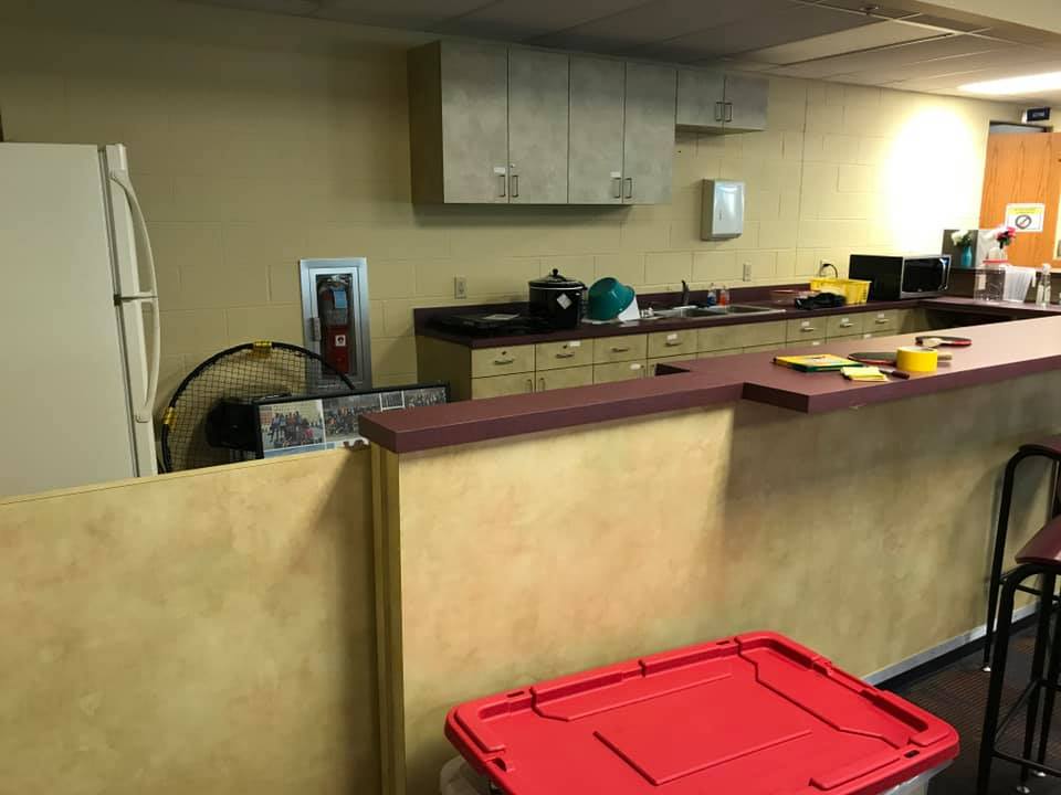 Small kitchen or break room with counters, cabinets, sink, and a red storage bin in the foreground.