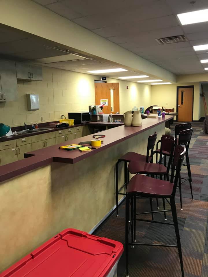 Kitchen or break room with long counter, stools, sinks, and red storage bin under fluorescent lighting