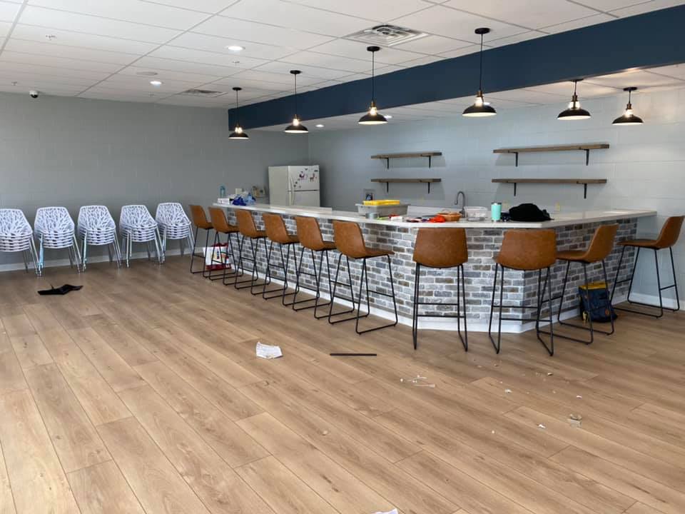 Empty event room with long tables, stools, pendant lights, and stacked white chairs on wood flooring.