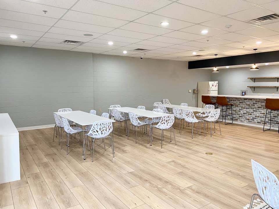 Empty modern event room with white tables and chairs, wood floor, and a bar area along the back wall