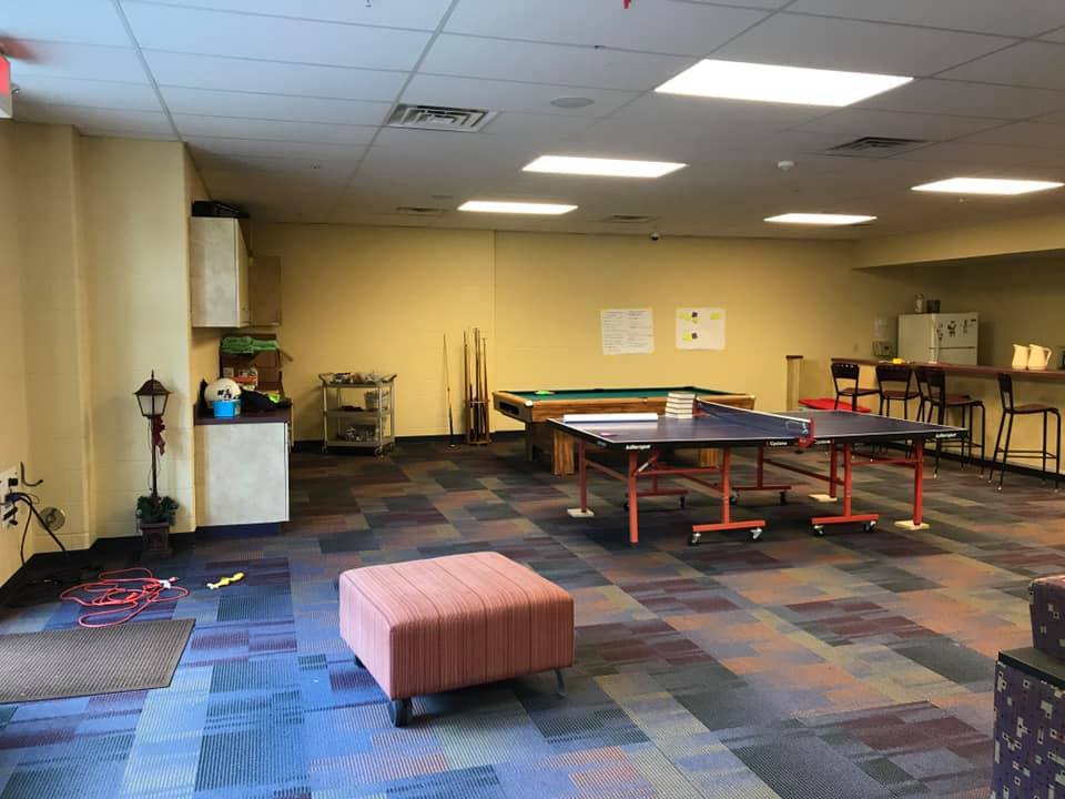 Recreation room with ping-pong tables, ottoman, and carpeted floor under fluorescent lights