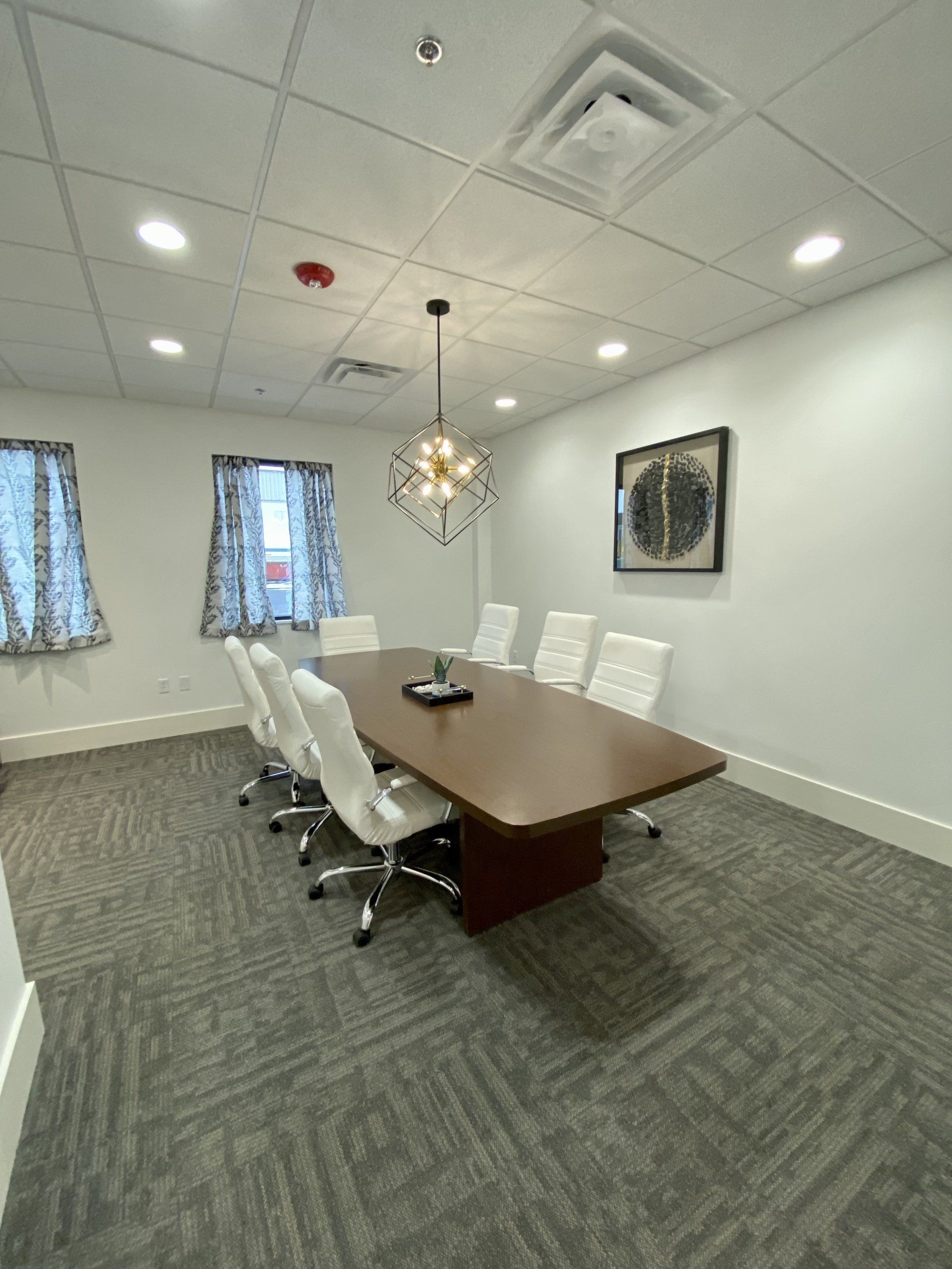 Empty conference room with white chairs, wooden table, patterned carpet, ceiling lights, and wall art.