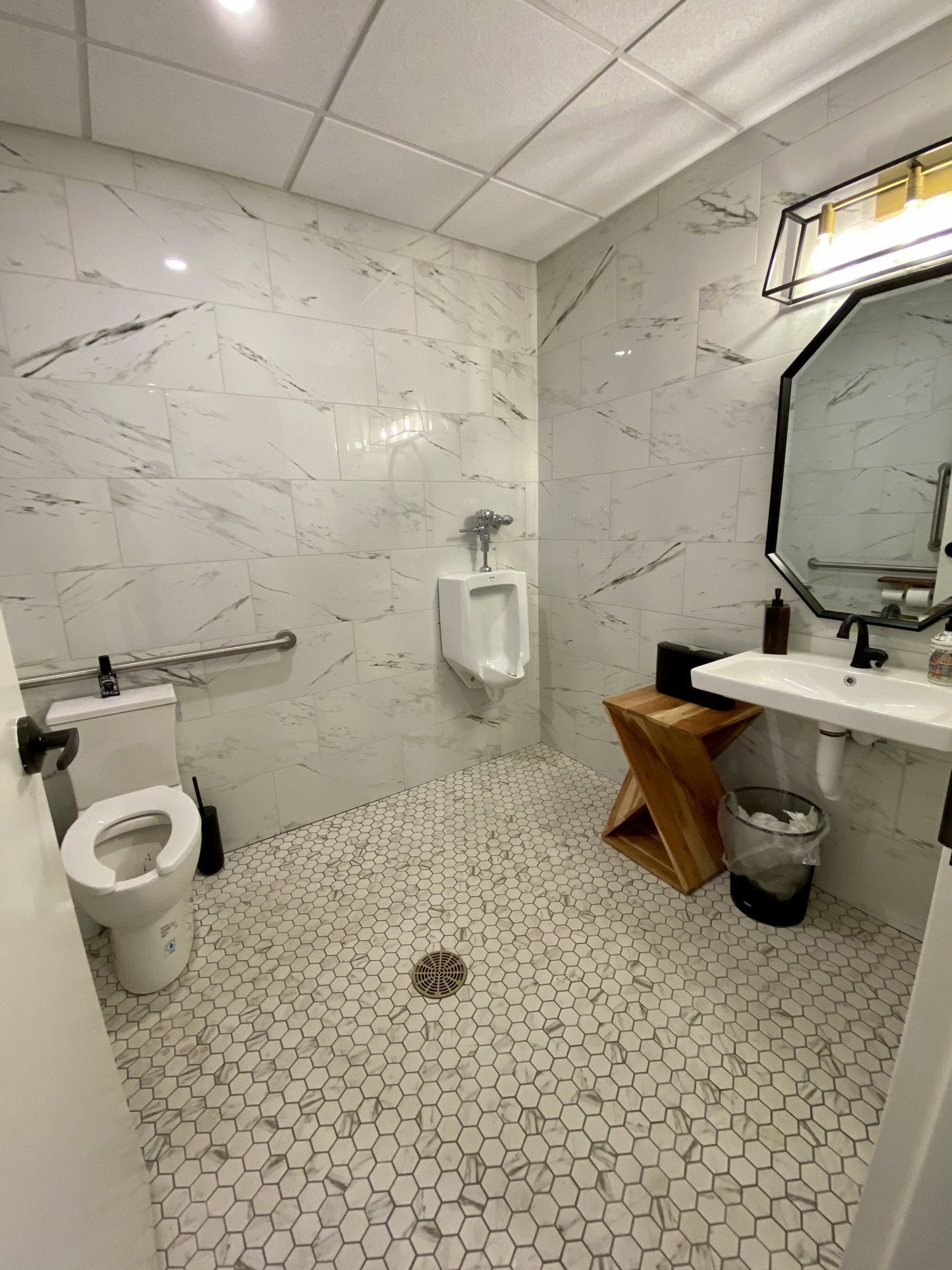 Small tiled public restroom with toilet, sink, mirror, urinal, and paper towel dispenser