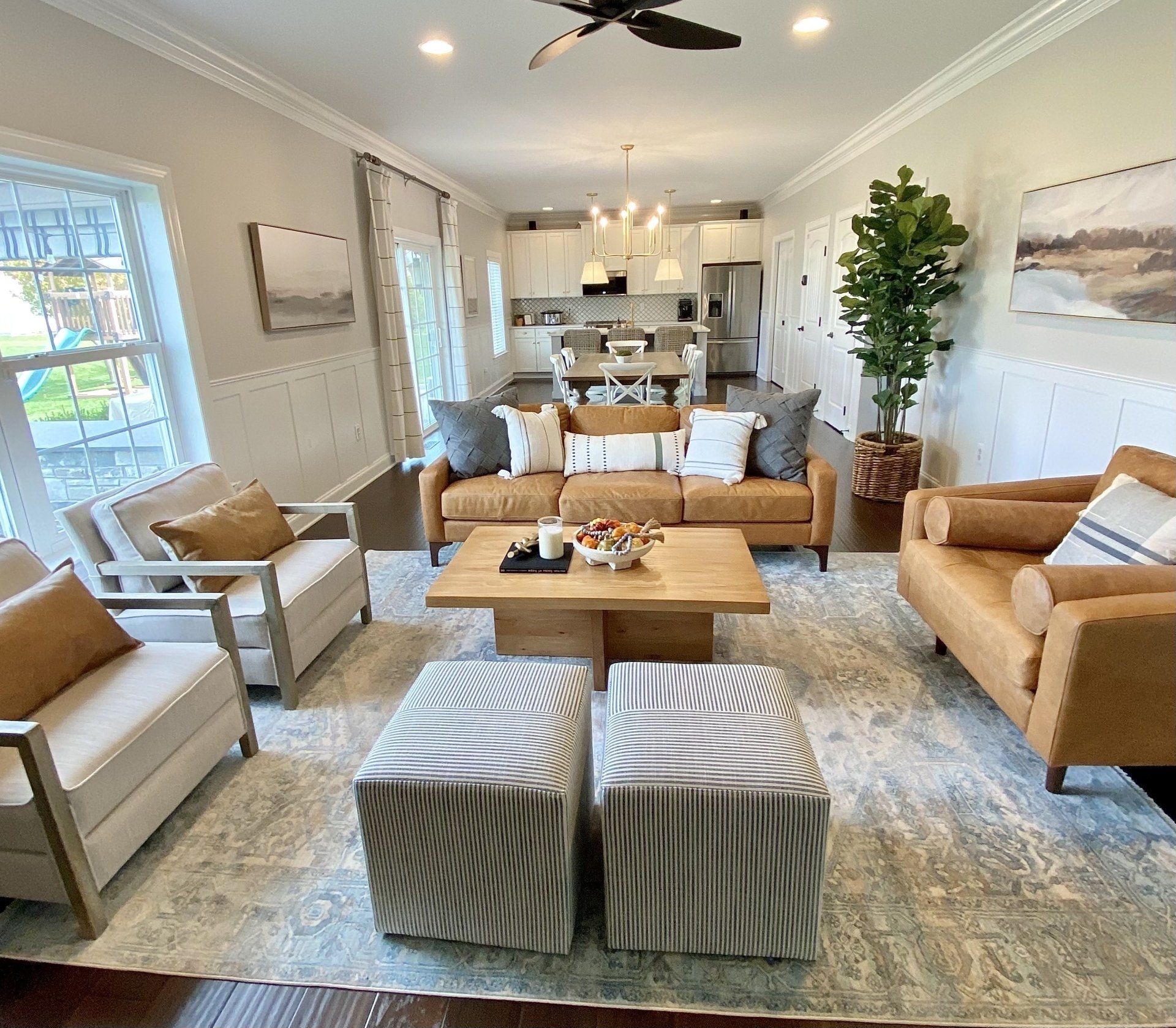 Bright modern living room with beige sofas, striped ottomans, and an open kitchen in the background
