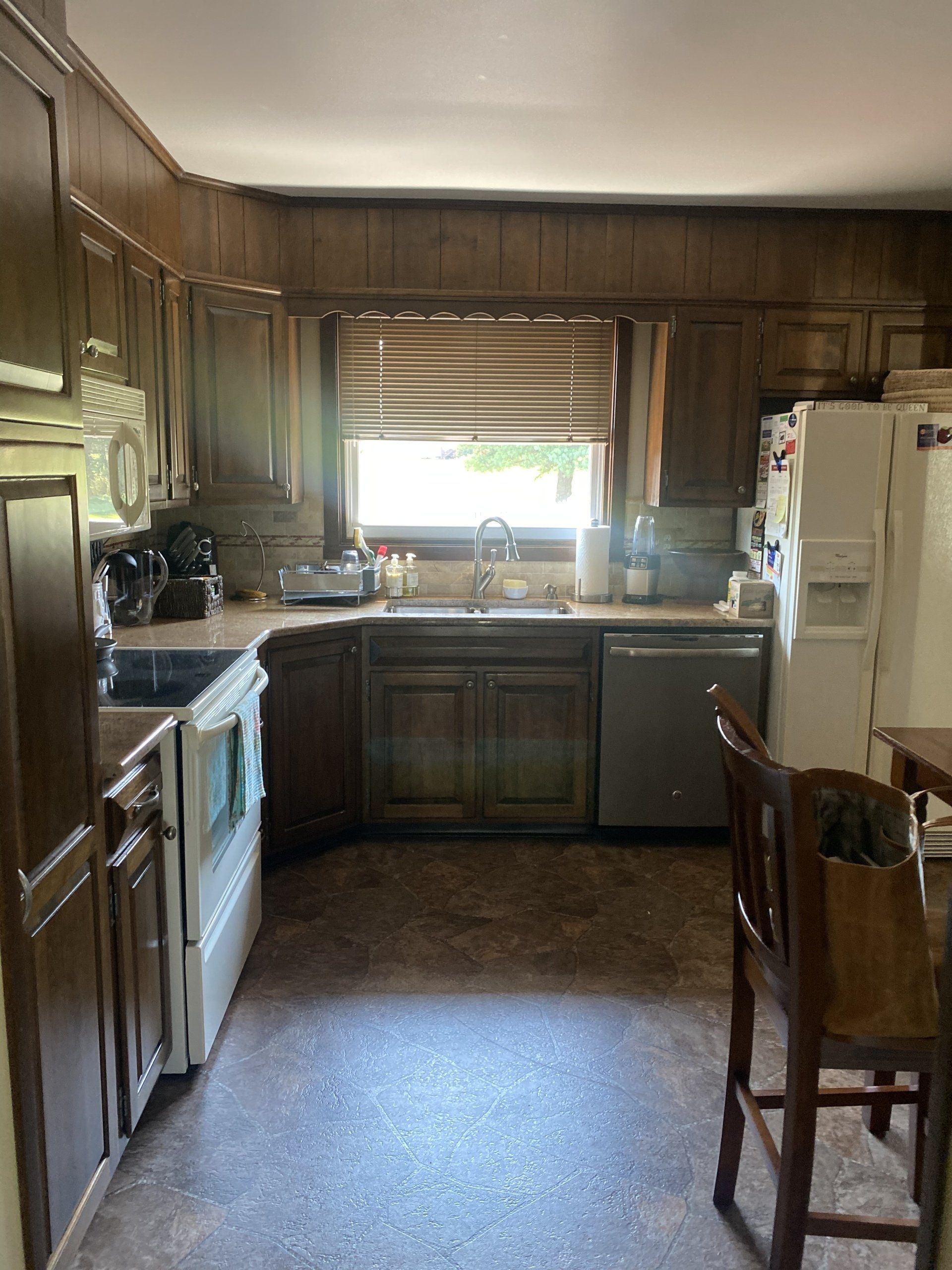 Dim wood-paneled kitchen with white appliances, sink under window, and a wooden chair in the foreground