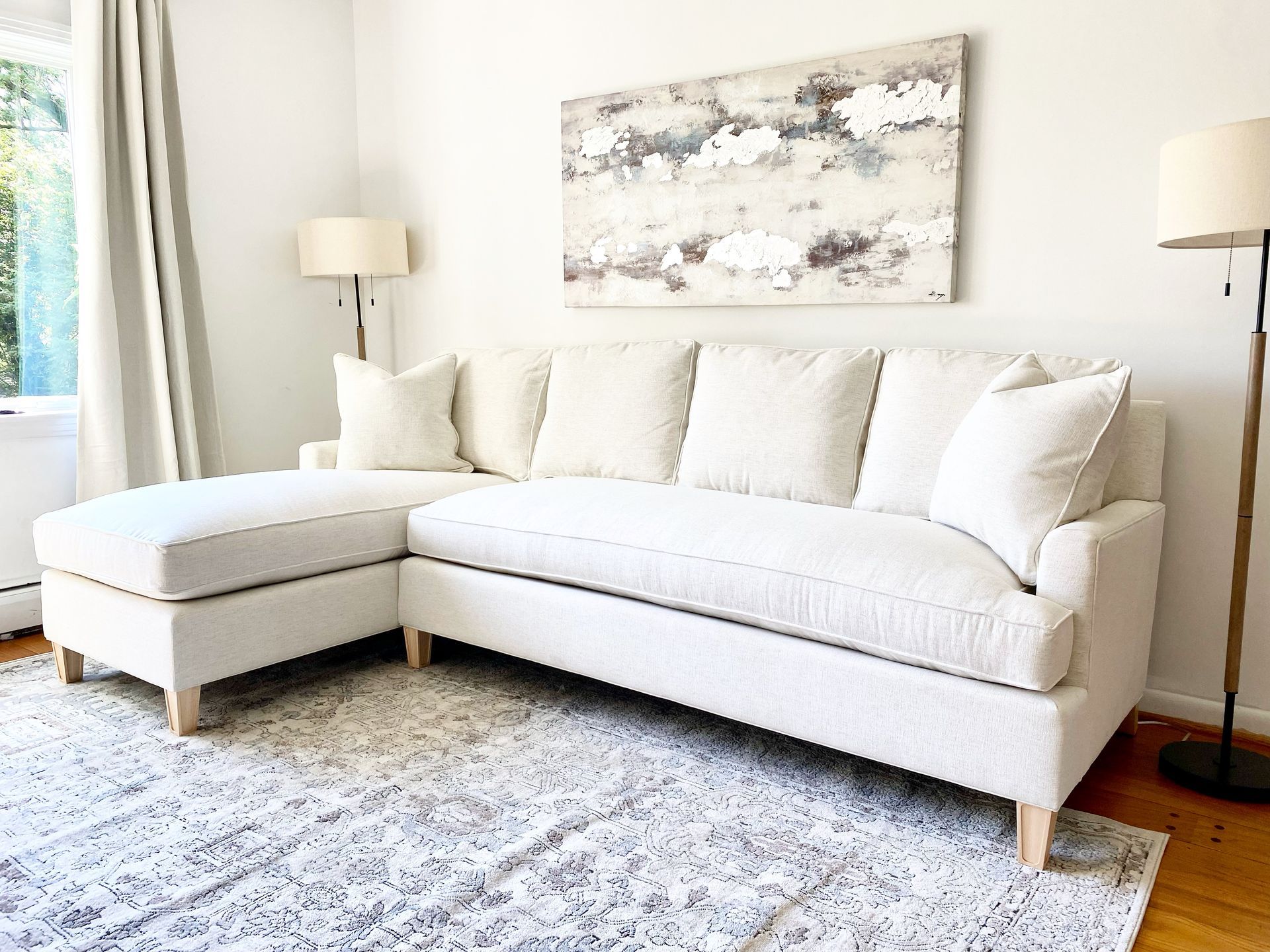 Bright living room with a white sectional sofa, matching ottoman, lamp, and abstract wall art