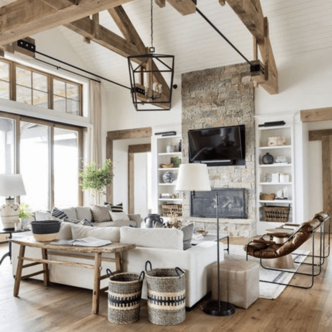 Bright rustic living room with stone fireplace, exposed beams, large windows, and neutral furniture
