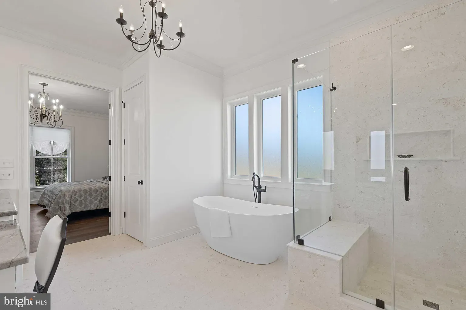Bright white bathroom with freestanding tub, glass shower, chandelier, and adjacent bedroom visible through open door