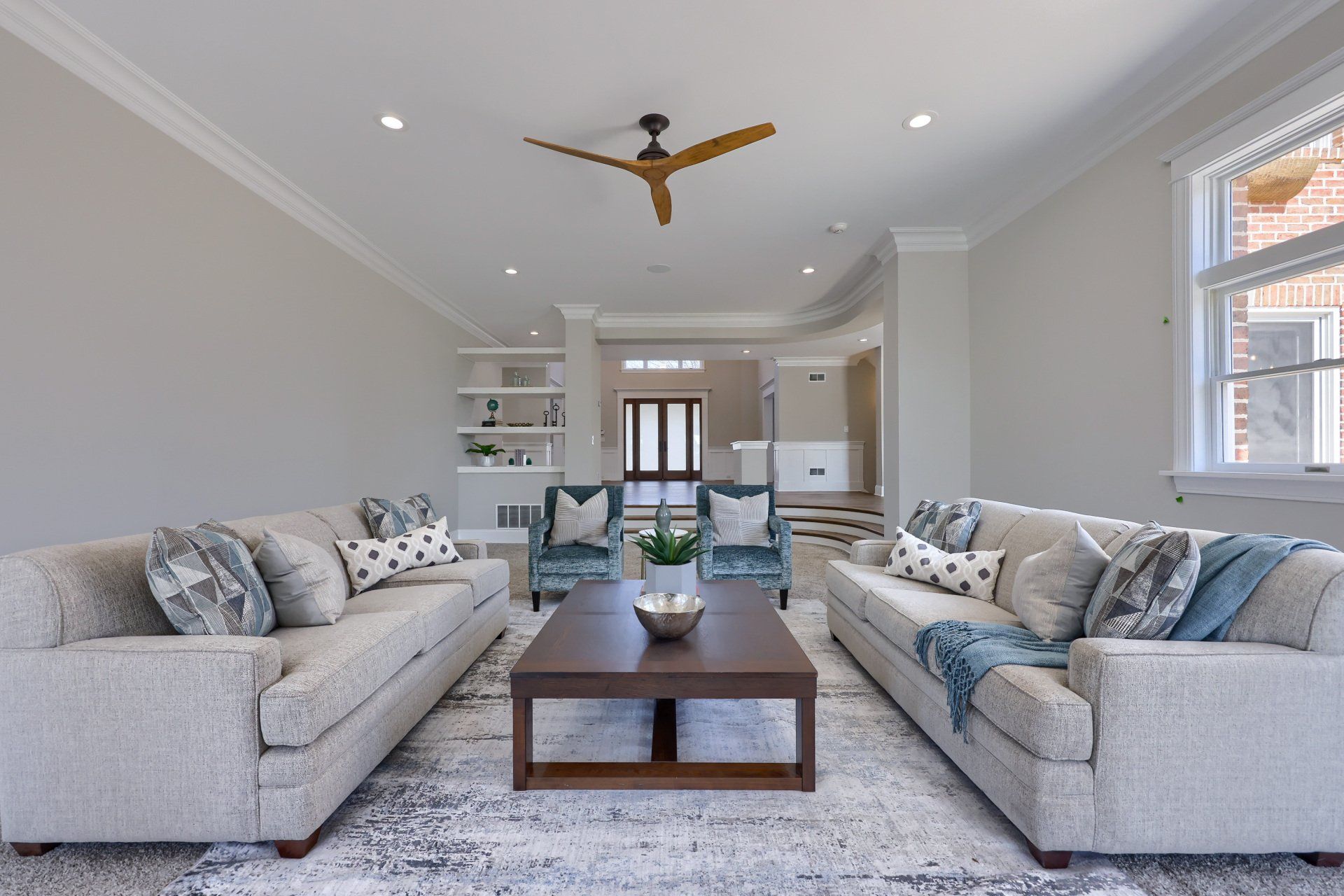 Bright modern living room with two gray sofas, coffee table, rug, ceiling fan, and open kitchen beyond