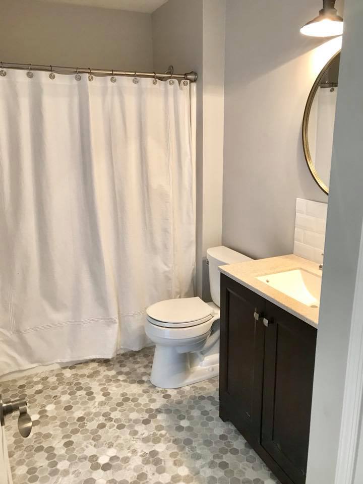 Small bathroom with white shower curtain, toilet, mosaic tile floor, and dark vanity with round mirror.