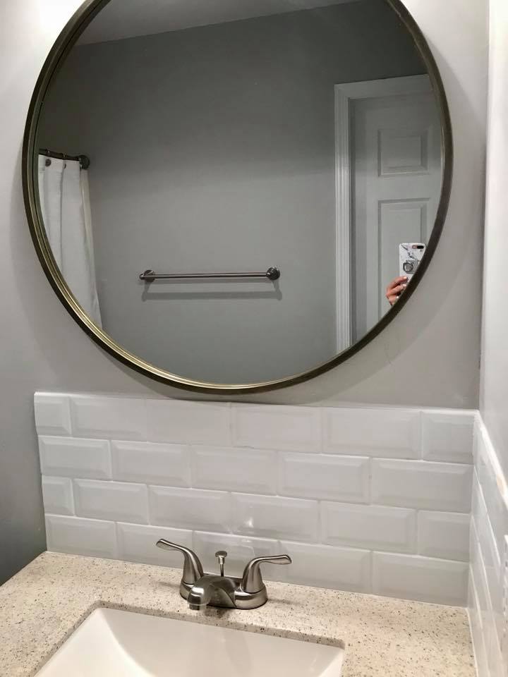 Bathroom vanity with round mirror, gray walls, white tile backsplash, and single sink faucet