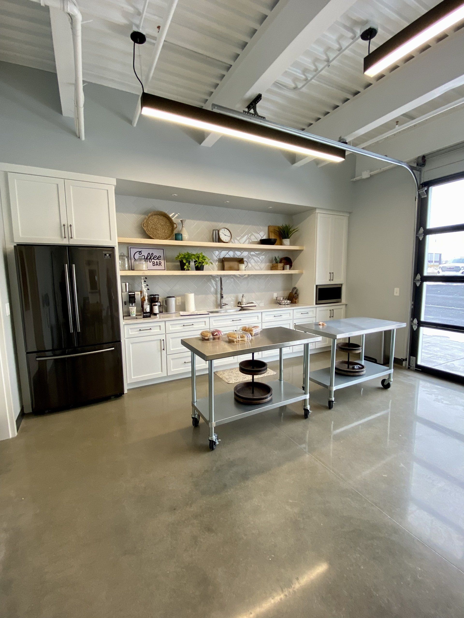 Bright industrial kitchen with white cabinets, black refrigerator, open shelves, and stainless prep tables.