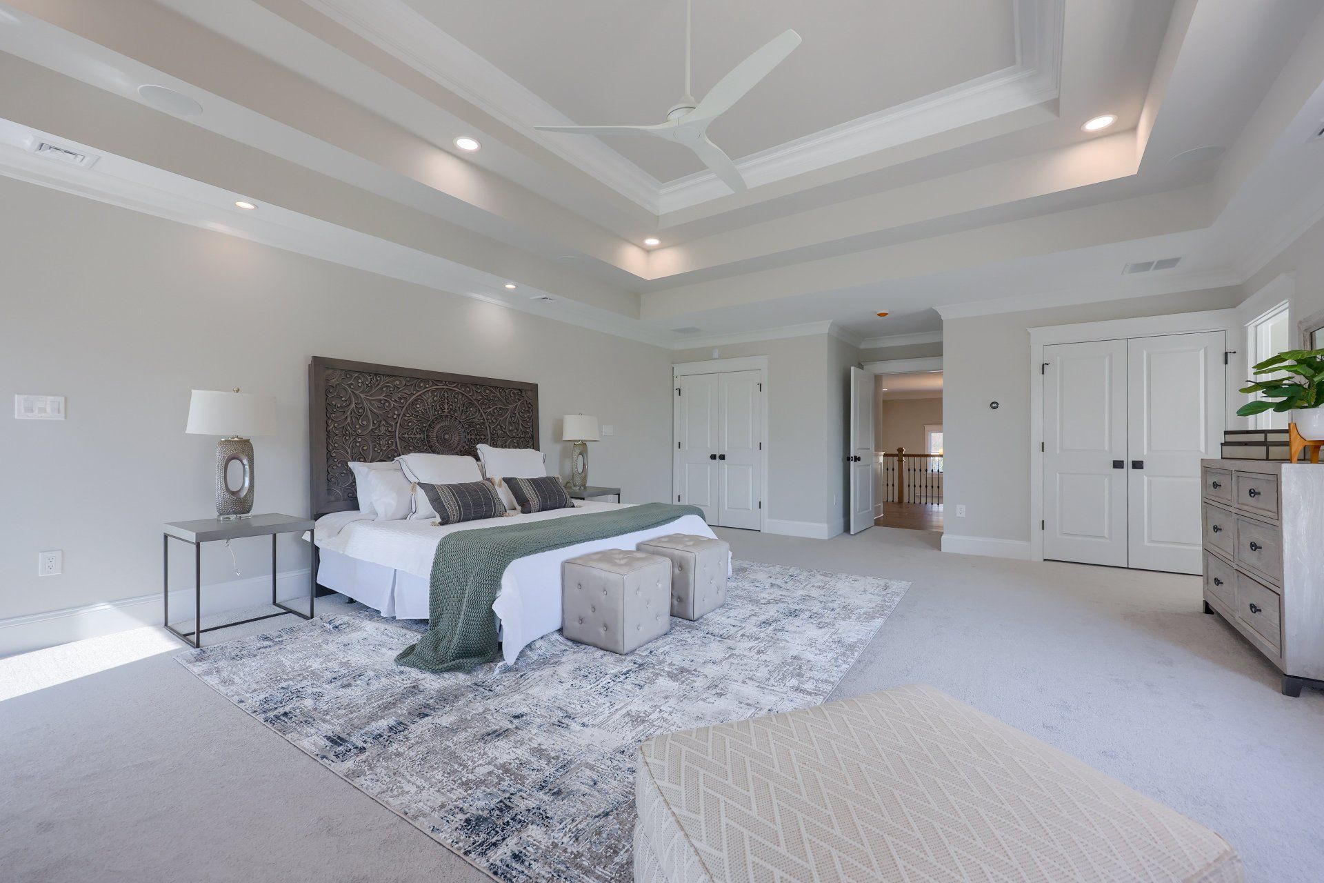 Bright modern bedroom with white walls, gray carpet, large bed, and tufted bench under a tray ceiling.