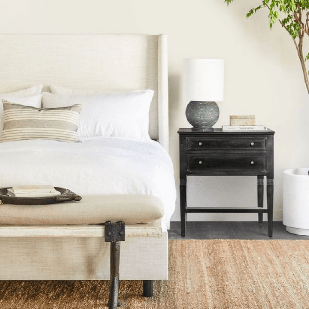 Minimal bedroom with beige bed, black nightstand, lamp, and woven rug.