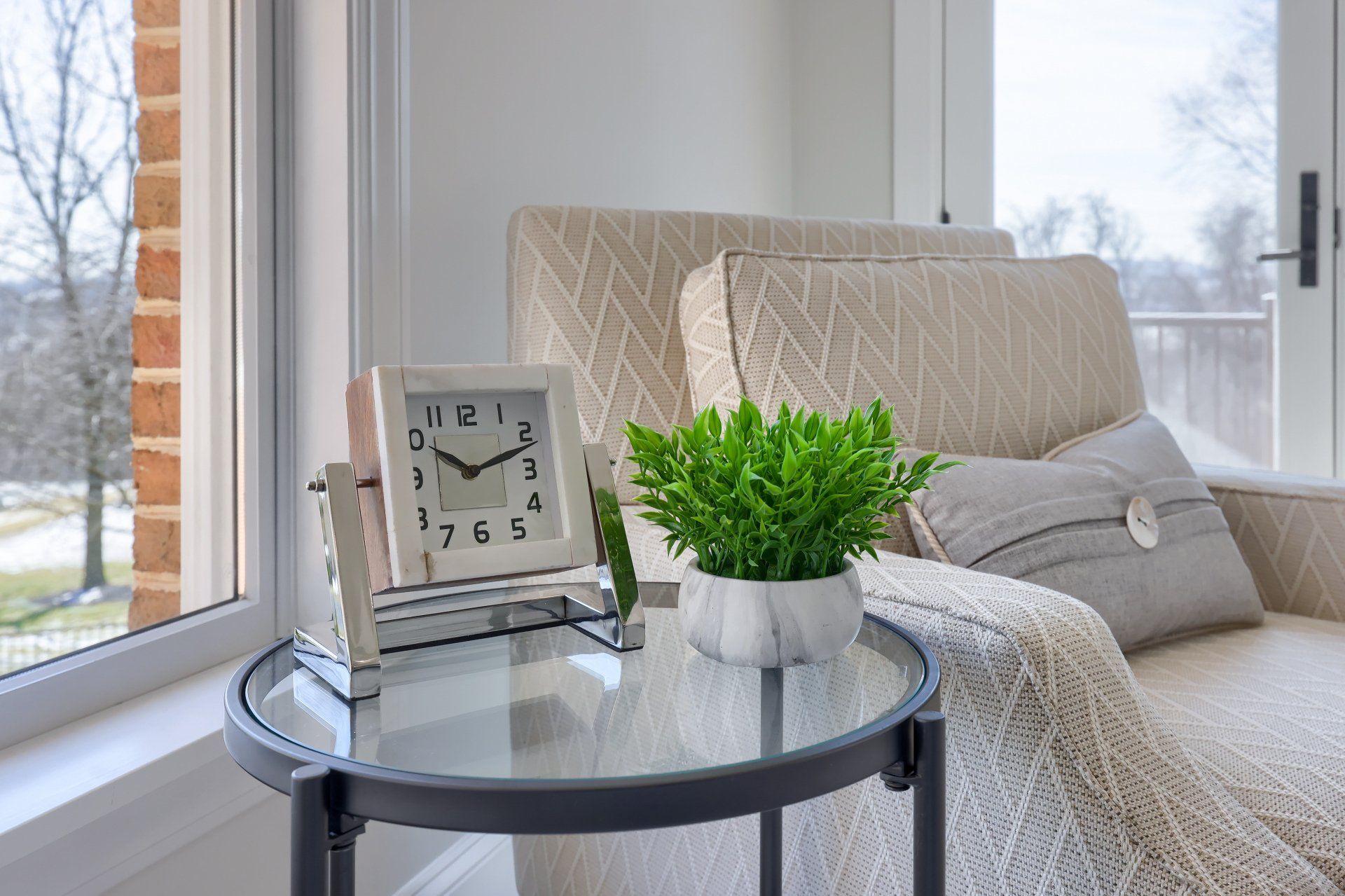 Cozy window nook with a beige chair, glass side table, clock, and potted plant by snowy windows.