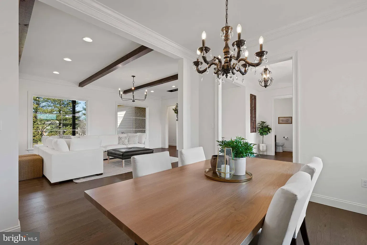 Bright modern dining room with wooden table, white chairs, chandelier, and open living area beyond.