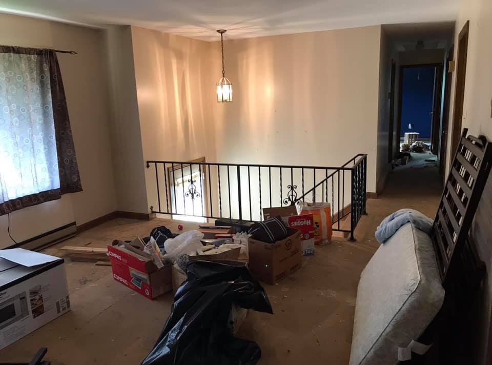 Cluttered upstairs landing with boxes, furniture, and a black railing beside a hallway window.
