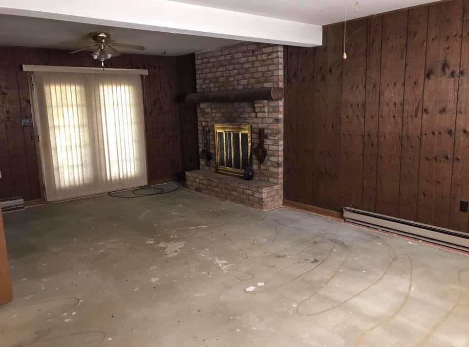 Empty living room with brick fireplace, wood-paneled wall, and large curtained window