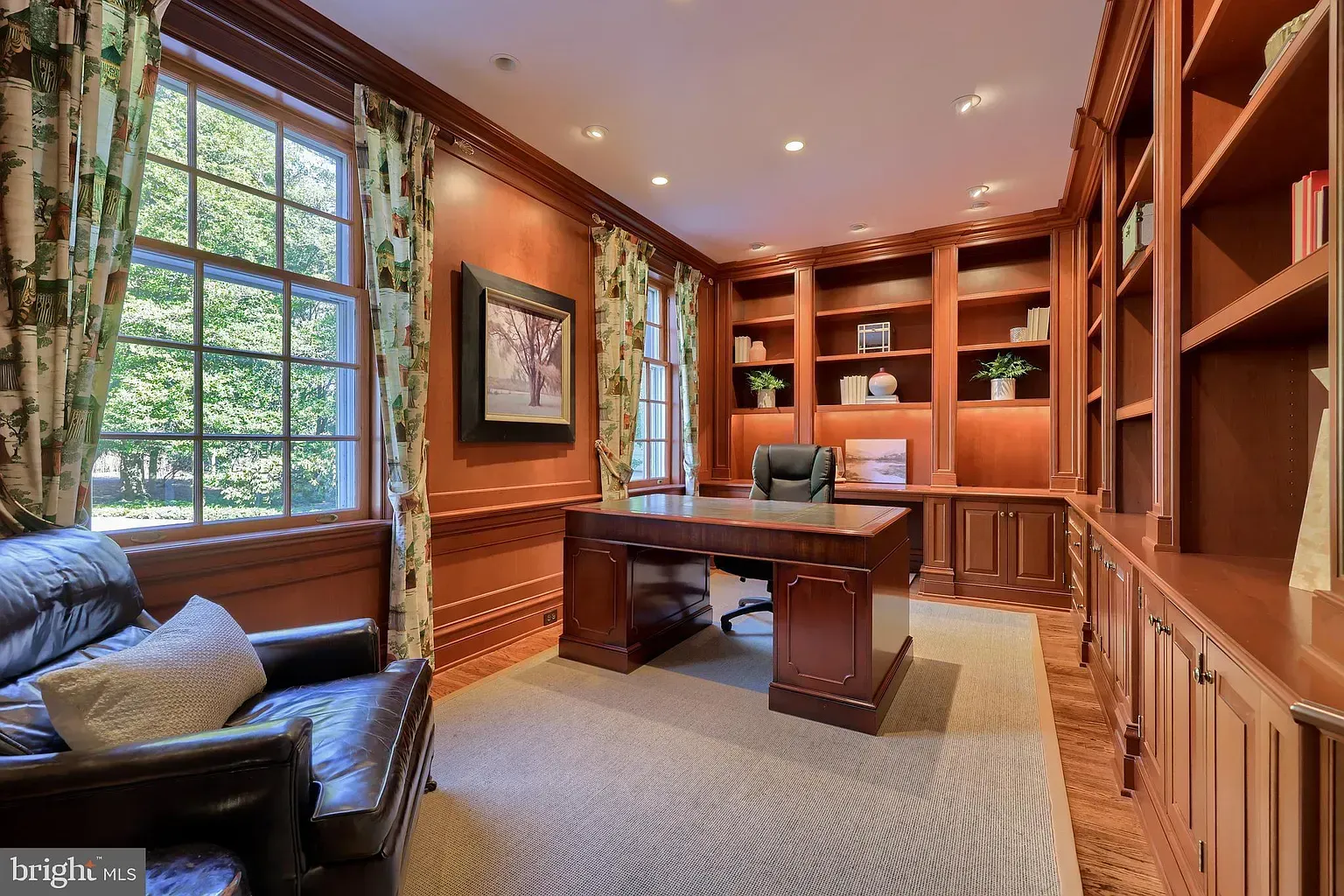 Elegant home office with wooden bookshelves, leather chair, desk, and large window with greenery.