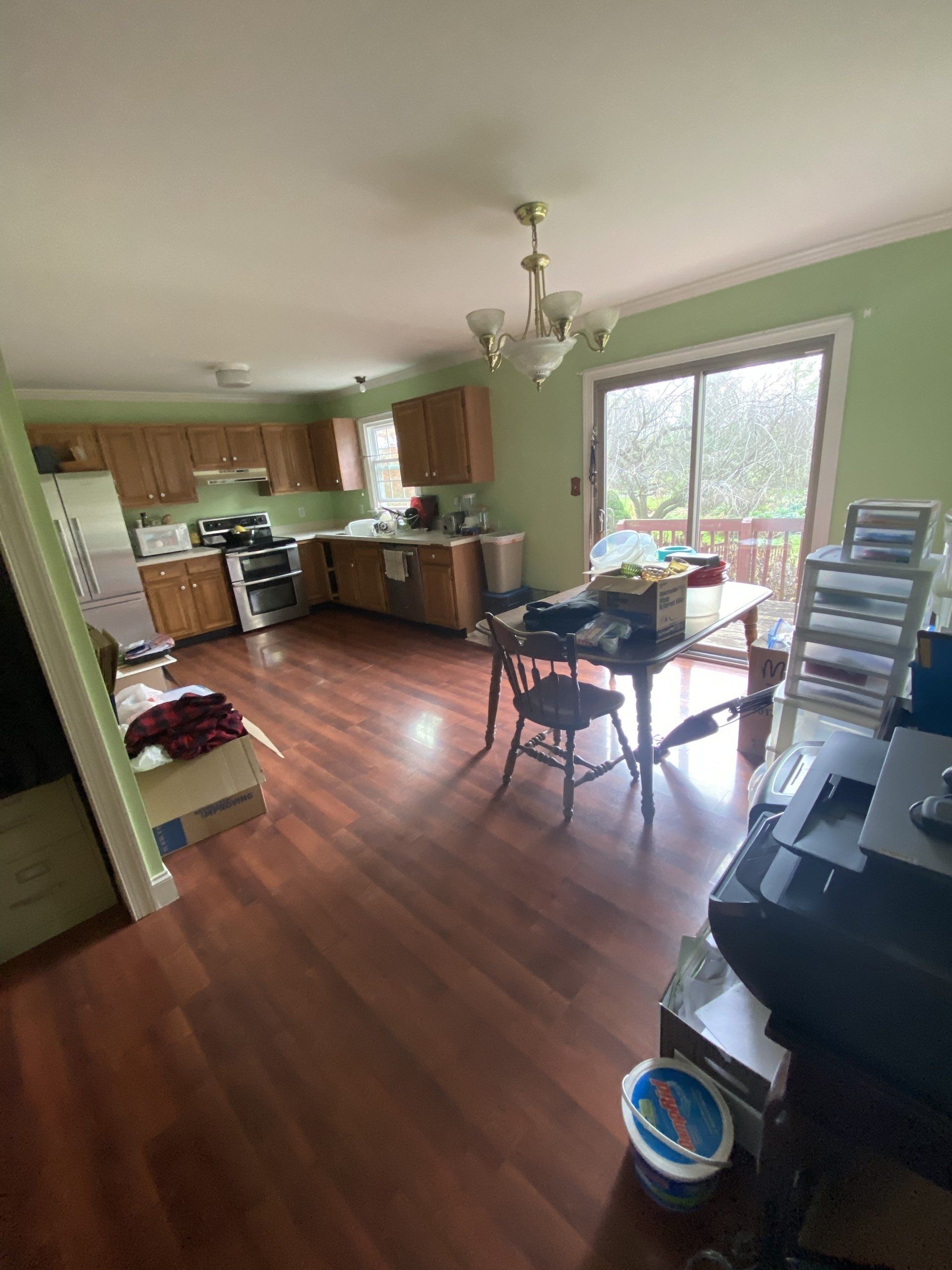Cluttered open-plan kitchen and dining area with wood floors, a table, chairs, and large windows.