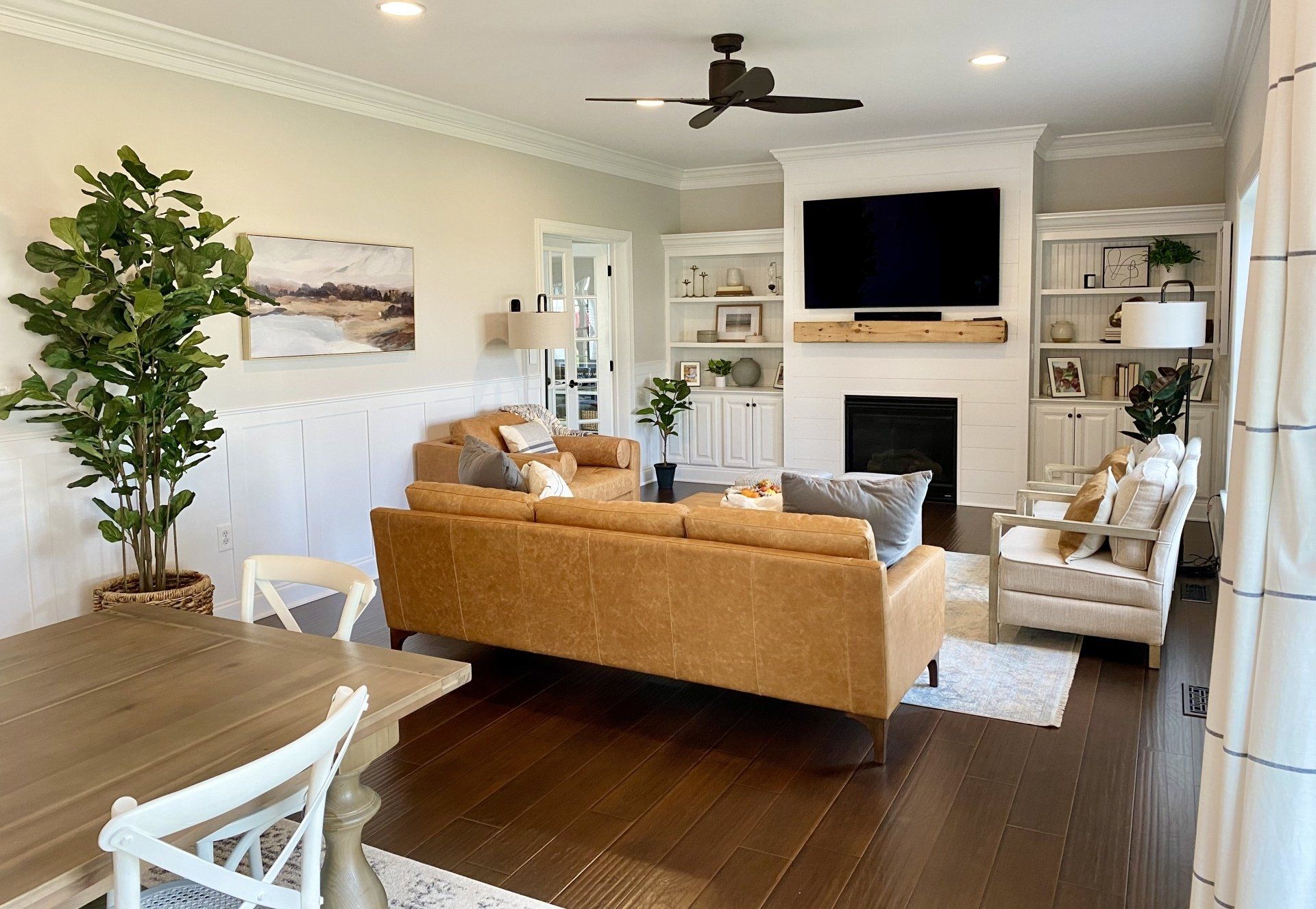 Bright living room with tan sofa, fireplace, wall TV, white chairs, and a large potted plant