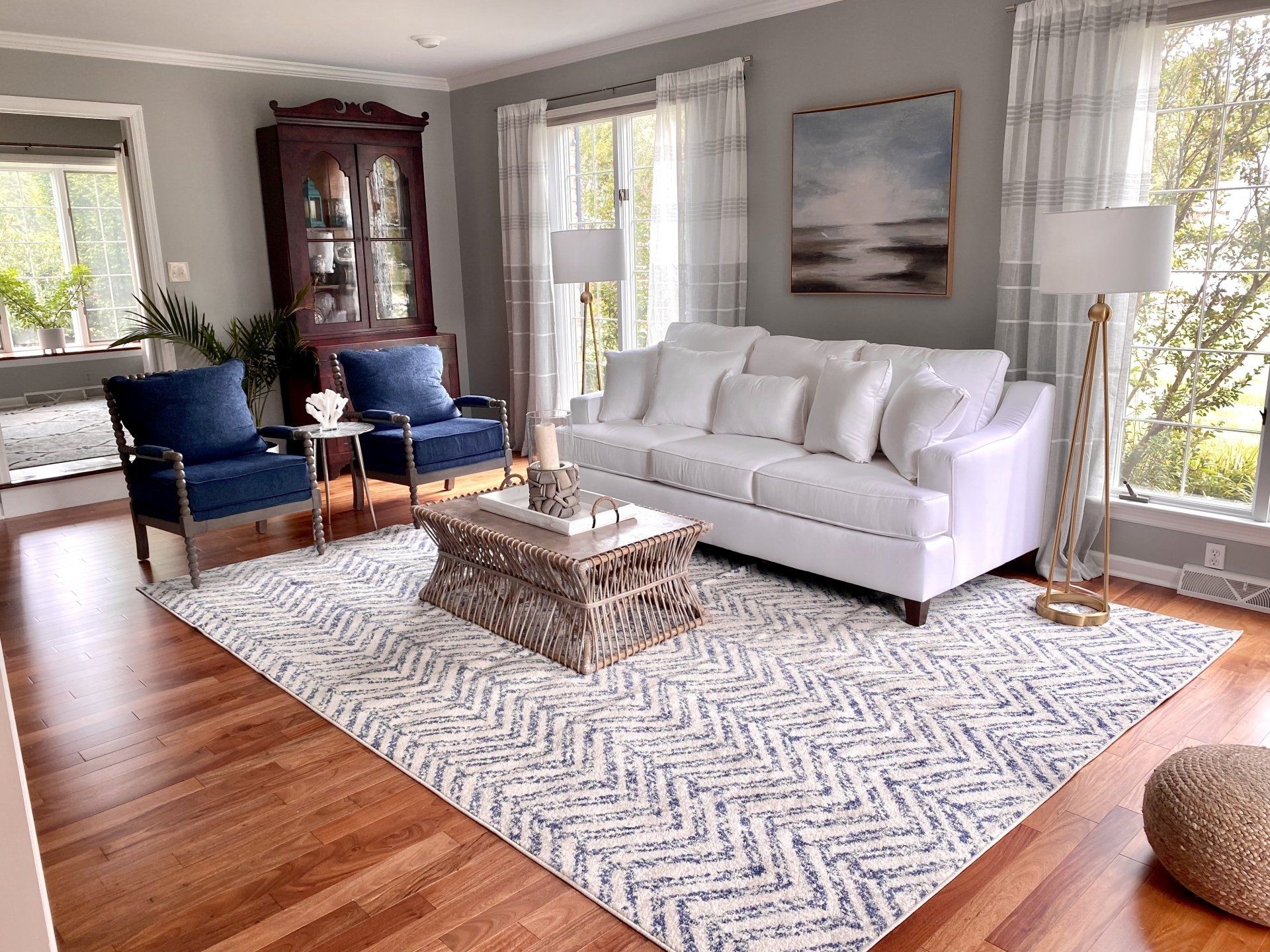 Bright living room with white sofa, blue chairs, patterned rug, and large windows showing trees