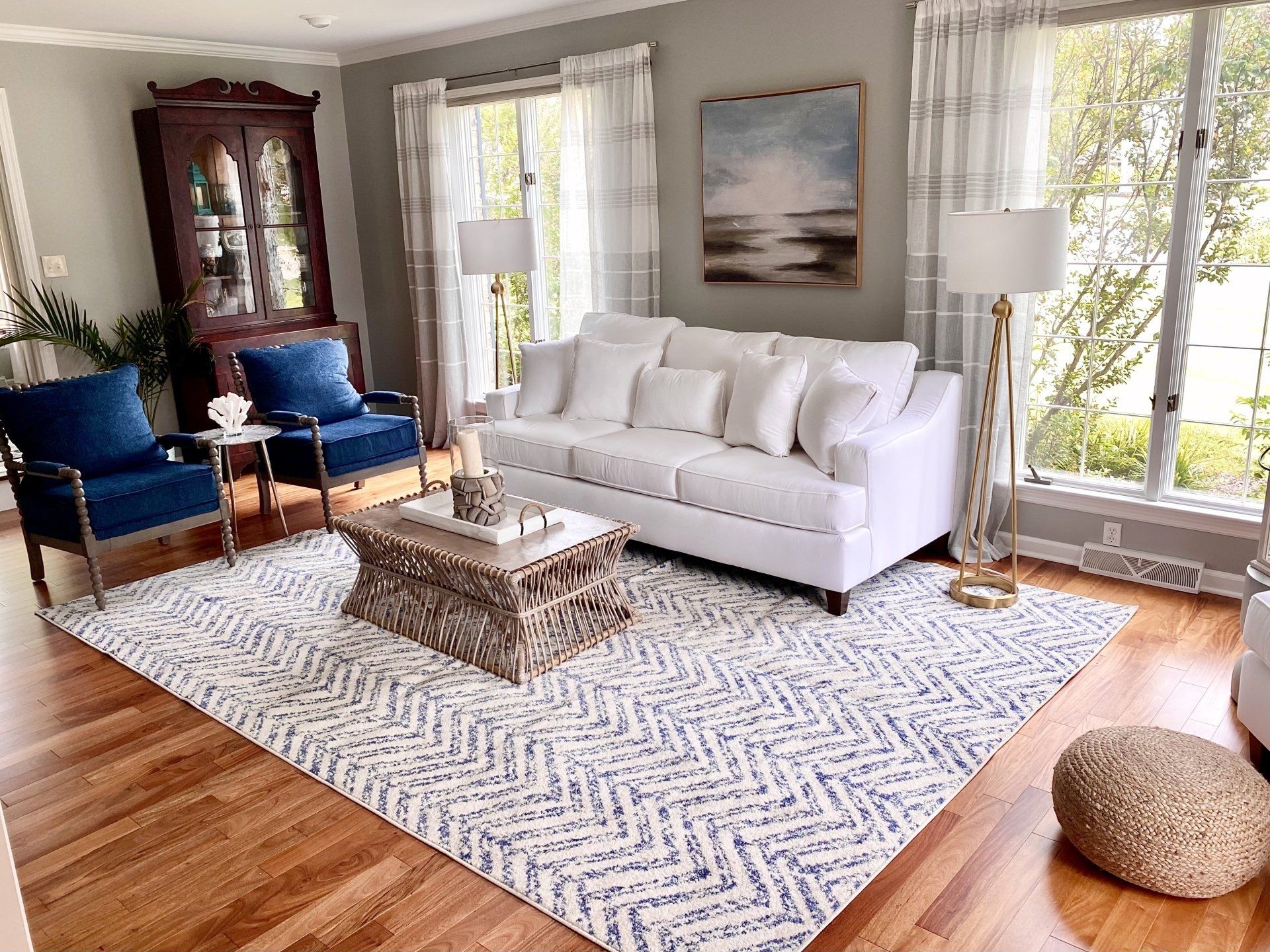 Bright living room with white sofa, blue chairs, coffee table, patterned rug, and large windows